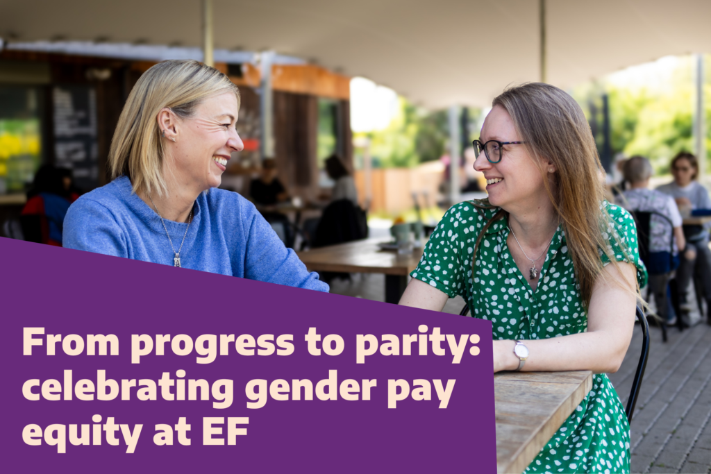 Two ladies sitting at a table in a coffee shop chatting and smiling. Text overlay reads, "From progress to parity: celebrating gender pay equity at EF
