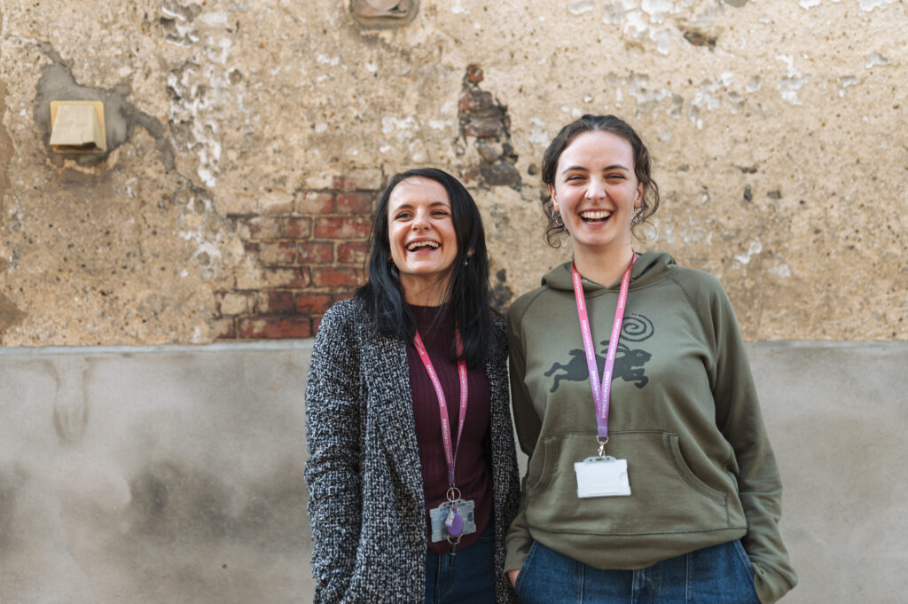 Two women against a stone wall laugh wearing Emerging Futures Lanyards