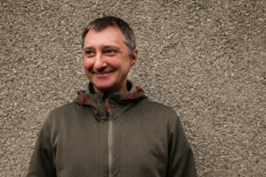 A person wearing an olive-green hooded jacket standing in front of a textured stone wall