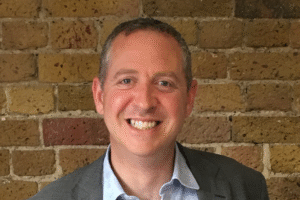 A person wearing a grey suit jacket and light blue shirt standing in front of a rustic brick wall