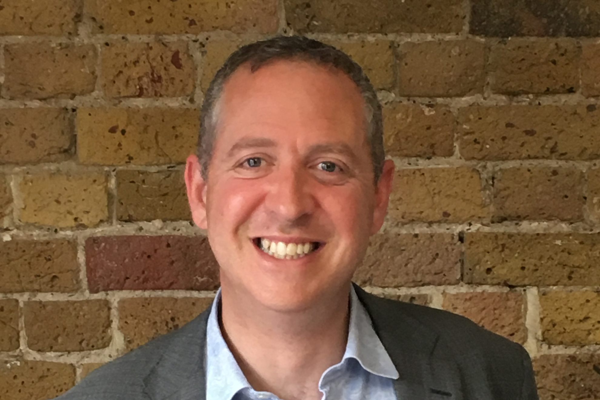 A person wearing a grey suit jacket and light blue shirt standing in front of a rustic brick wall