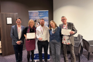 A group of five people standing together indoors, holding certificates and posing in front of a banner that reads “Career Matters – Lived Experience Charter.” The group is dressed in smart clothing, and the setting appears to be a conference or event space with chairs and a carpeted floor.
