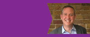A person in a grey suit jacket and light blue shirt standing in front of a brick wall, with a purple graphic background on the left side of the image