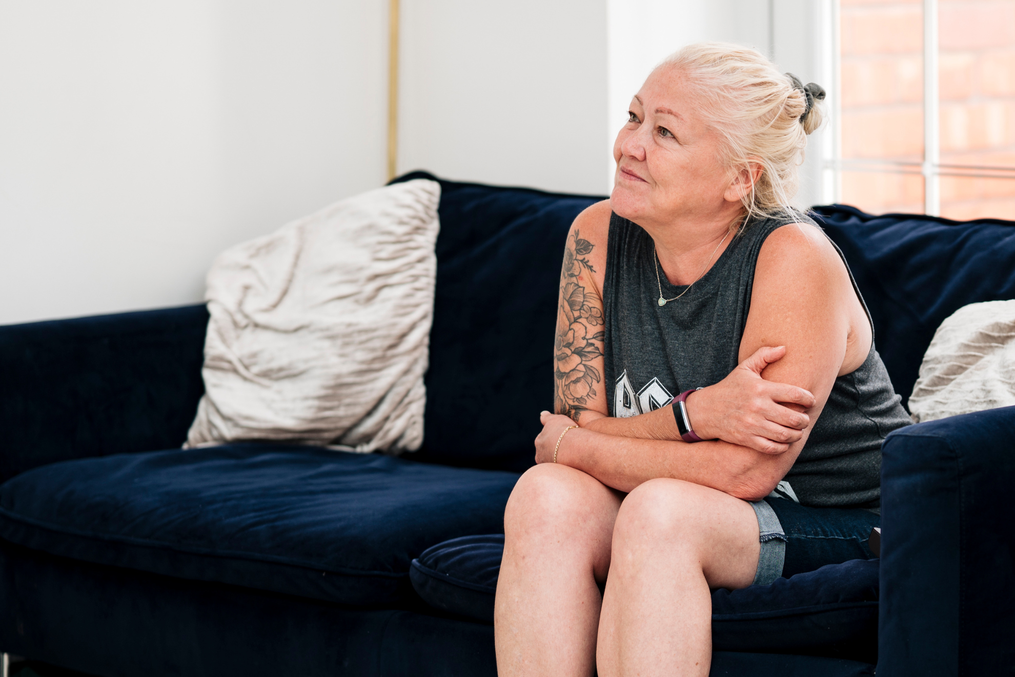 A person with light hair tied back sits on a dark blue sofa, arms folded, wearing a sleeveless top and shorts. A tattoo is visible on their arm, and light from a nearby window brightens the room.