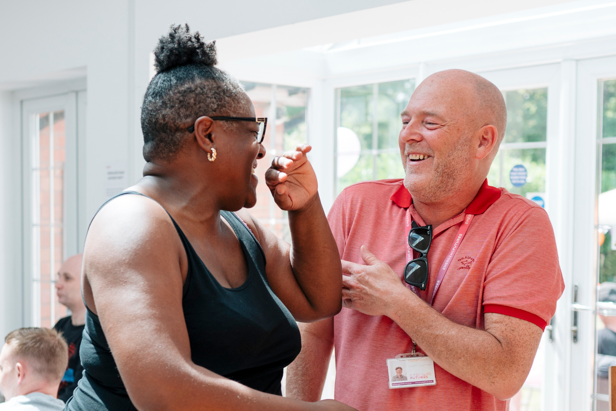 Two people standing indoors, facing each other and interacting in conversation. One person gestures with their hand while the other wears a lanyard and sunglasses, with bright windows in the background.