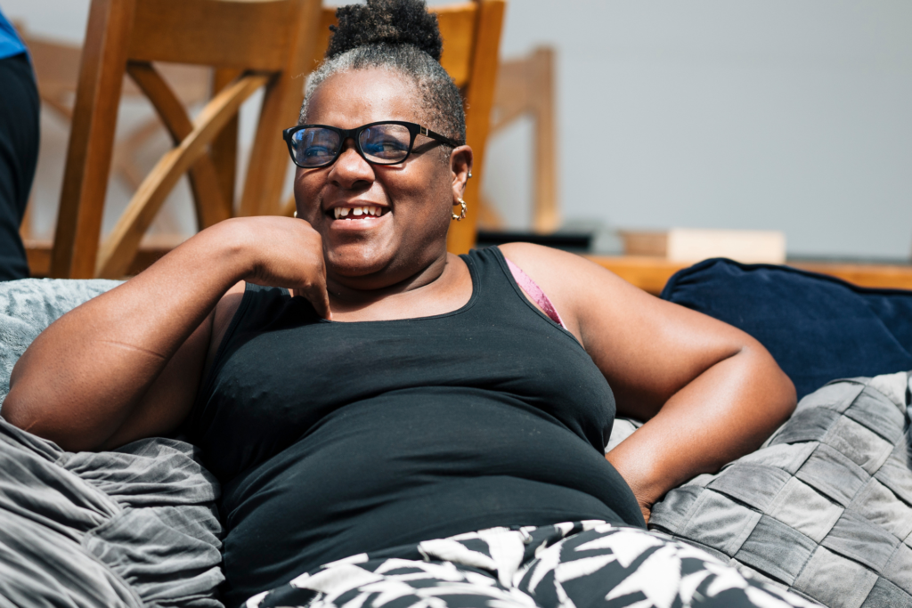 Person wearing glasses and a black sleeveless top sitting on a grey sofa, leaning back with one arm resting on the armrest. Wooden chairs are visible in the background of a bright indoor room.