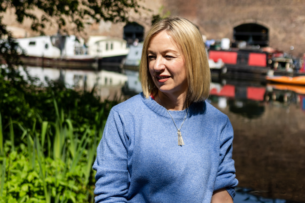 Person in a light blue jumper standing by a canal, with narrowboats and a brick building in the background.
