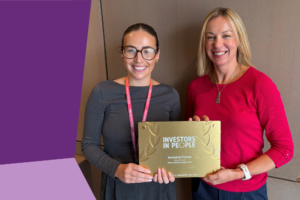 Two people stand indoors holding a gold Investors in People award plaque. One person is wearing glasses, a dark grey top and an Emerging Futures lanyard, while the other is wearing a bright pink jumper. The plaque displays the text "Investors in People" and recognises Emerging Futures for achieving We Invest in People Gold. A purple graphic panel appears along the left side of the image, with a neutral wood-panelled background behind the pair.