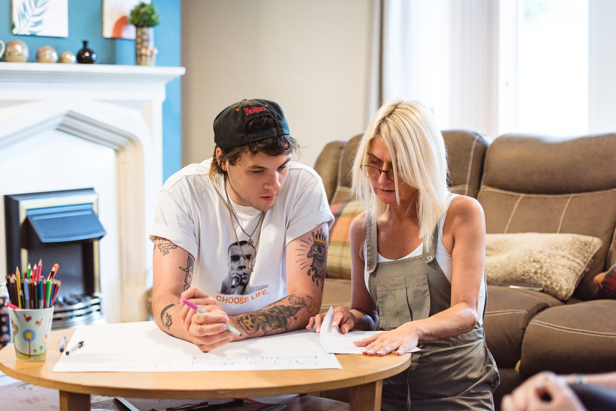 Two people sit together at a low table in a living room, reviewing paperwork and writing on large sheets of paper. One person holds a coloured pen while the other points to documents on the table. Coloured pencils and additional pens are visible nearby, with a sofa, fireplace and home furnishings in the background. The scene appears to show collaborative planning, learning or support activity in a relaxed, residential setting.