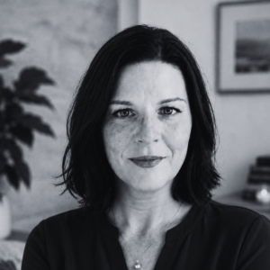 Black-and-white head-and-shoulders portrait of a person with shoulder-length dark hair, looking directly at the camera, with a softly blurred indoor background.