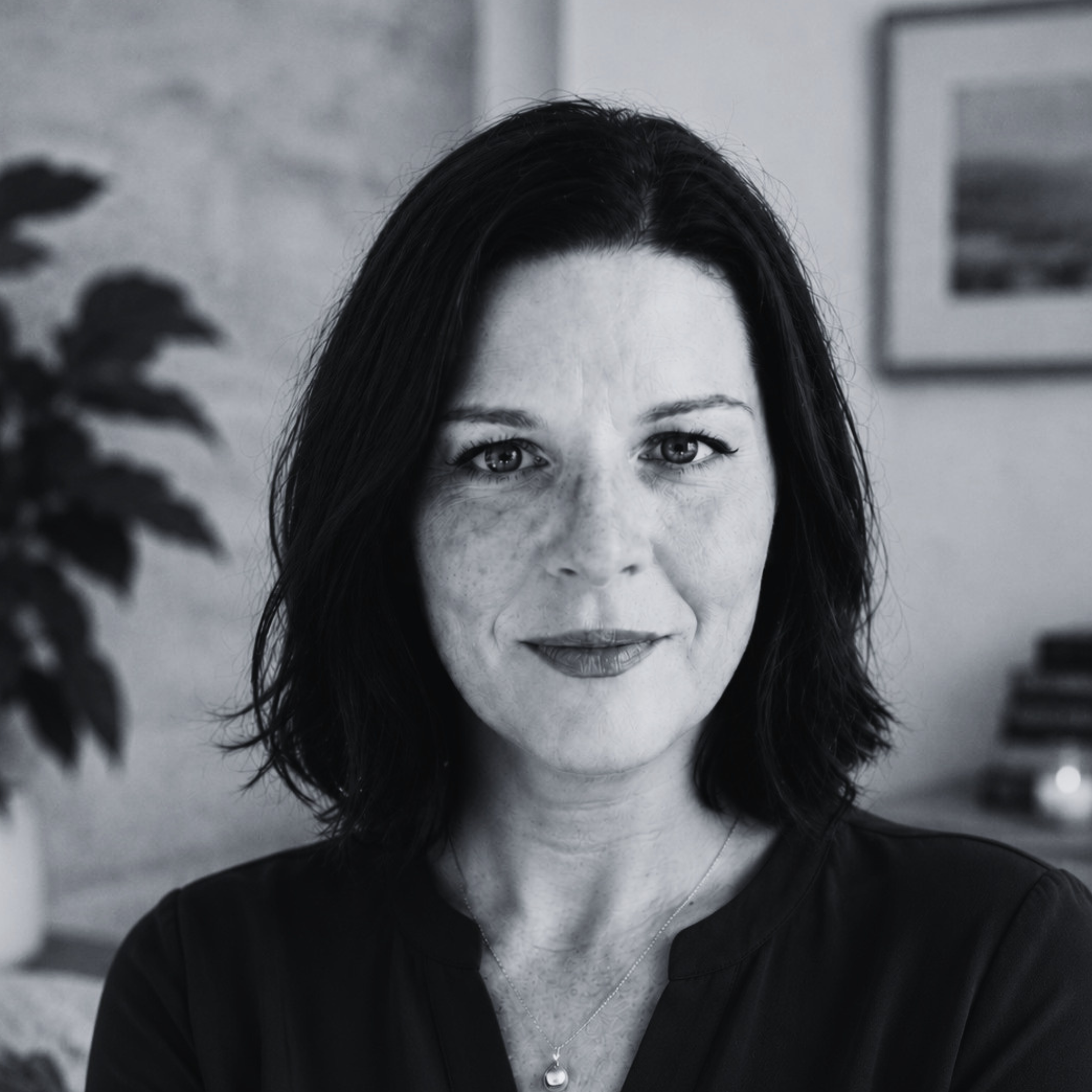 Black-and-white head-and-shoulders portrait of a person with shoulder-length dark hair, looking directly at the camera, with a softly blurred indoor background.