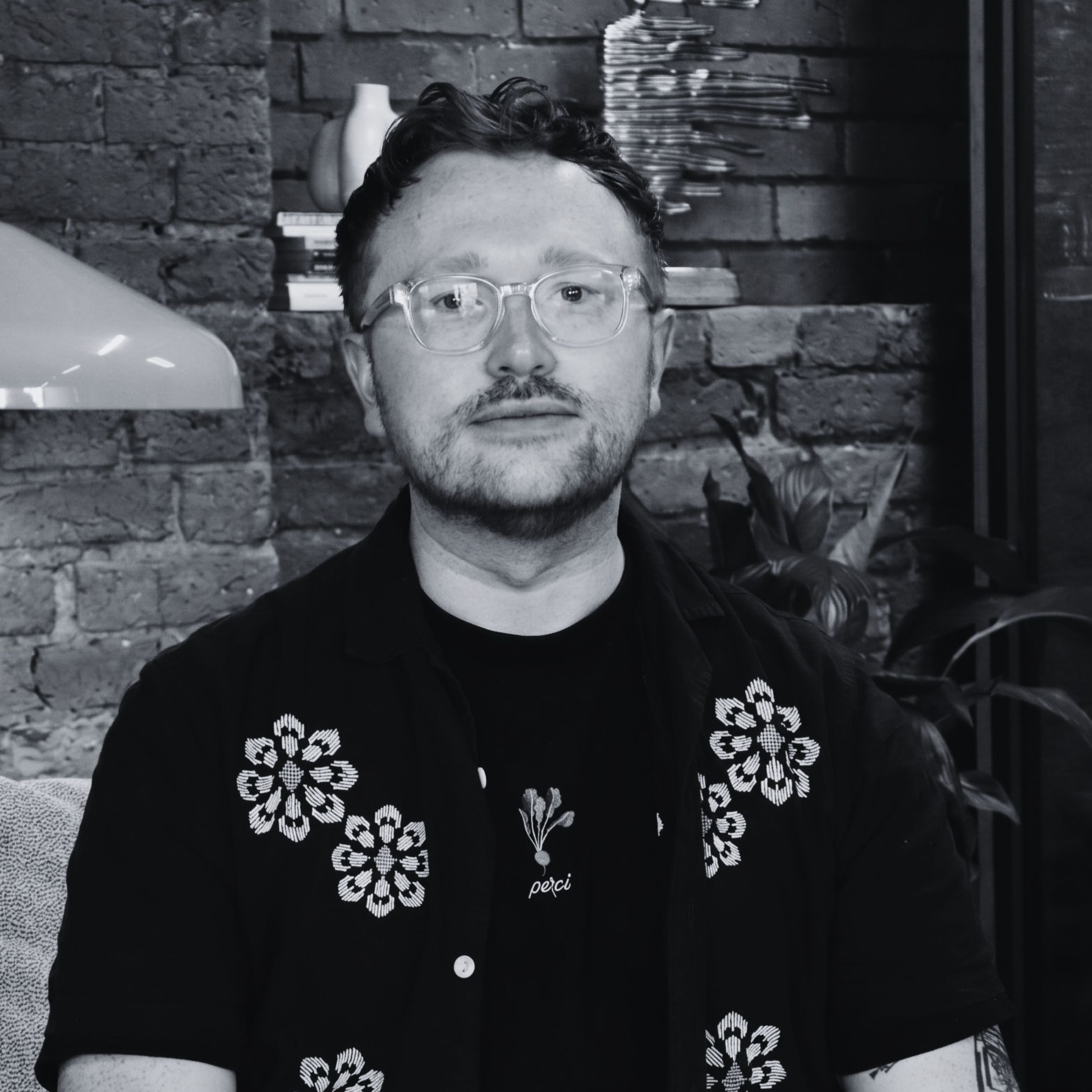 Black-and-white portrait of a person wearing glasses and a dark shirt with embroidered floral patterns, seated in front of an exposed brick wall.