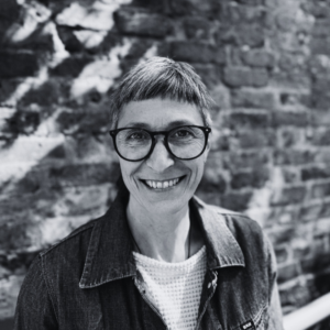 Black-and-white portrait of a person wearing glasses and a denim jacket, standing in front of a brick wall.
